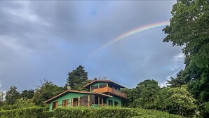 A two-story house with a balcony, surrounded by lush greenery and a rainbow in the sky. - Casa Campo Verde (Monteverde)