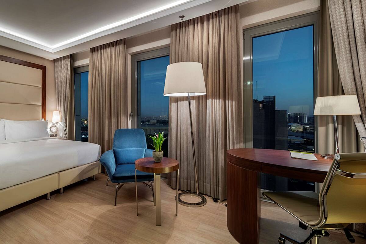 Quarto de hotel com uma cama grande, uma escrivaninha, uma cadeira, uma luminária e vista para a cidade à noite.