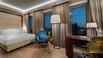 A hotel room with a large bed, a desk, a chair, a lamp, and a view of the city at night.