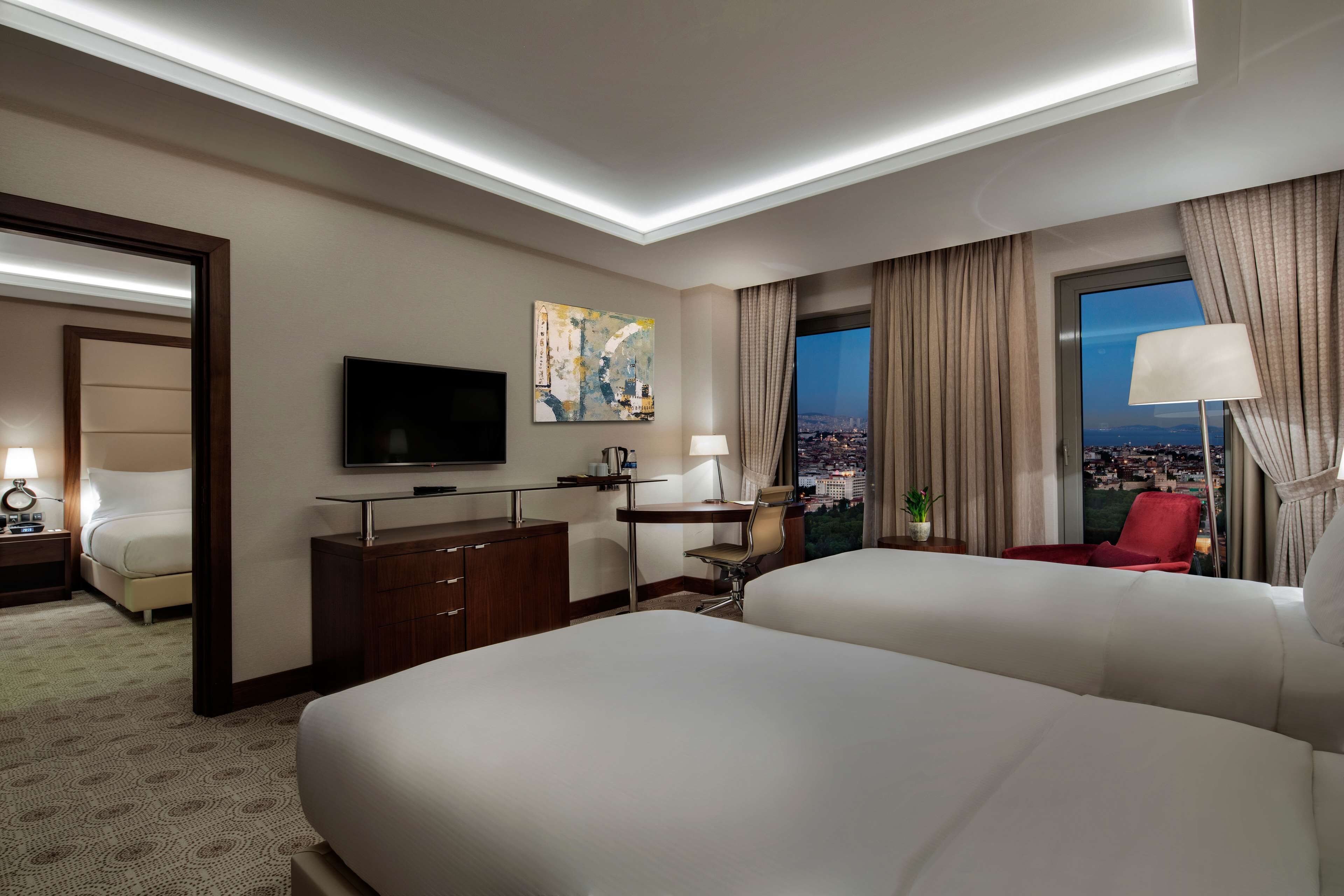 A hotel room with two beds, a TV, a desk, a chair, a lamp, and a window with a city view.