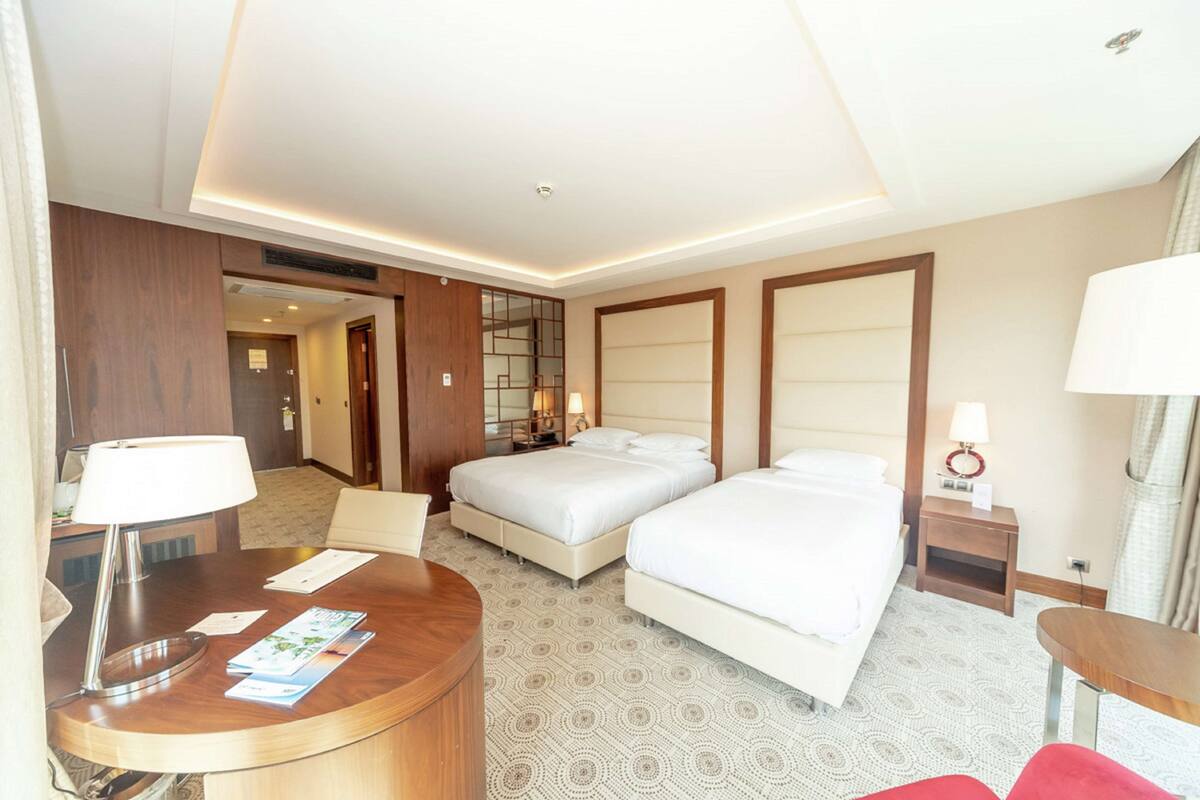 Quarto de hotel com duas camas, uma escrivaninha e uma mesinha.