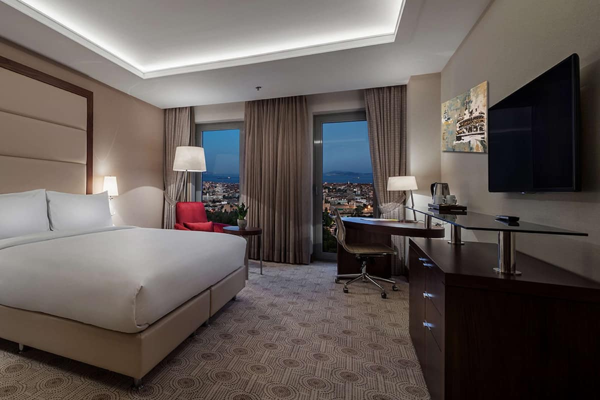 Quarto de hotel com uma cama grande, uma mesa de trabalho, televisão e vista para a cidade.