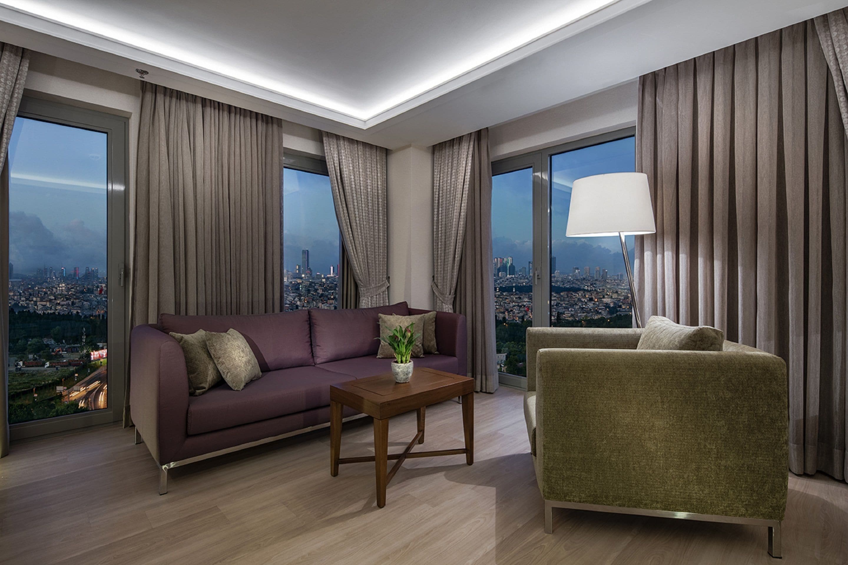A modern living room with a sofa, armchair, and coffee table, large windows with curtains, and a city view.