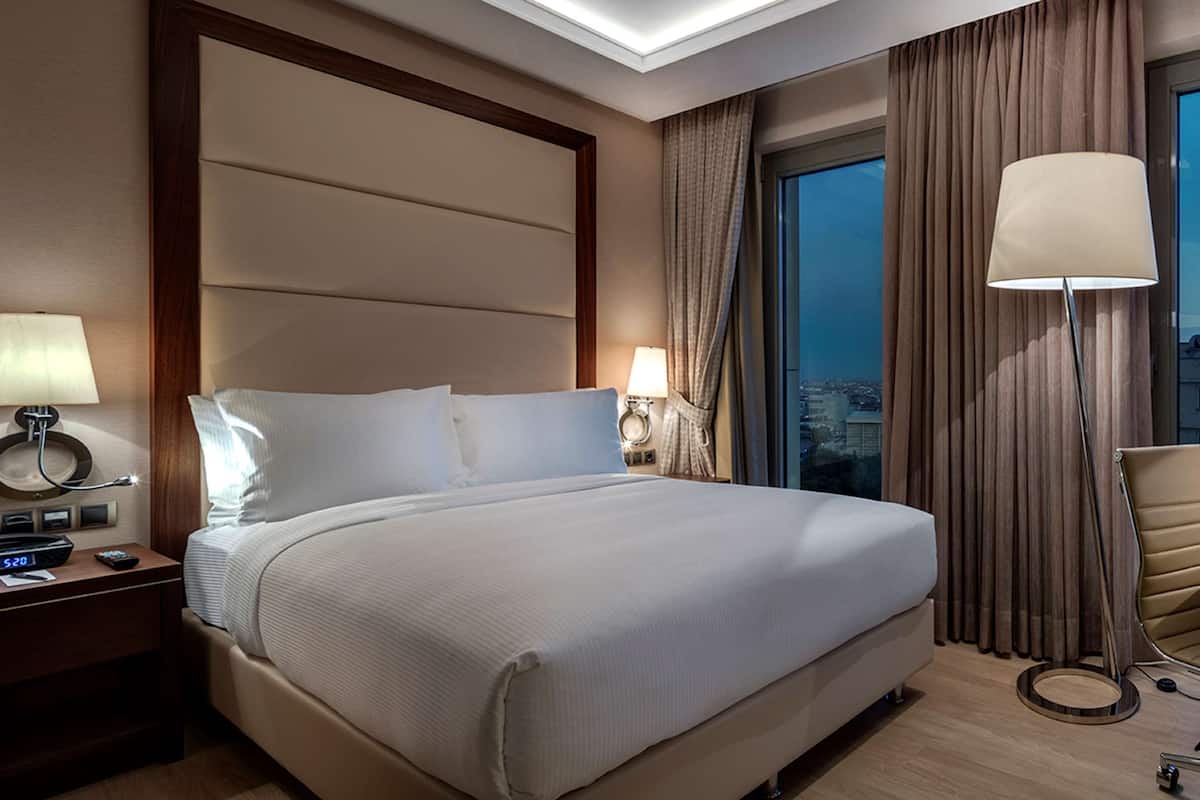 Quarto de hotel com uma cama grande, duas luminárias de cabeceira, uma escrivaninha com cadeira e vista para a cidade pela janela.