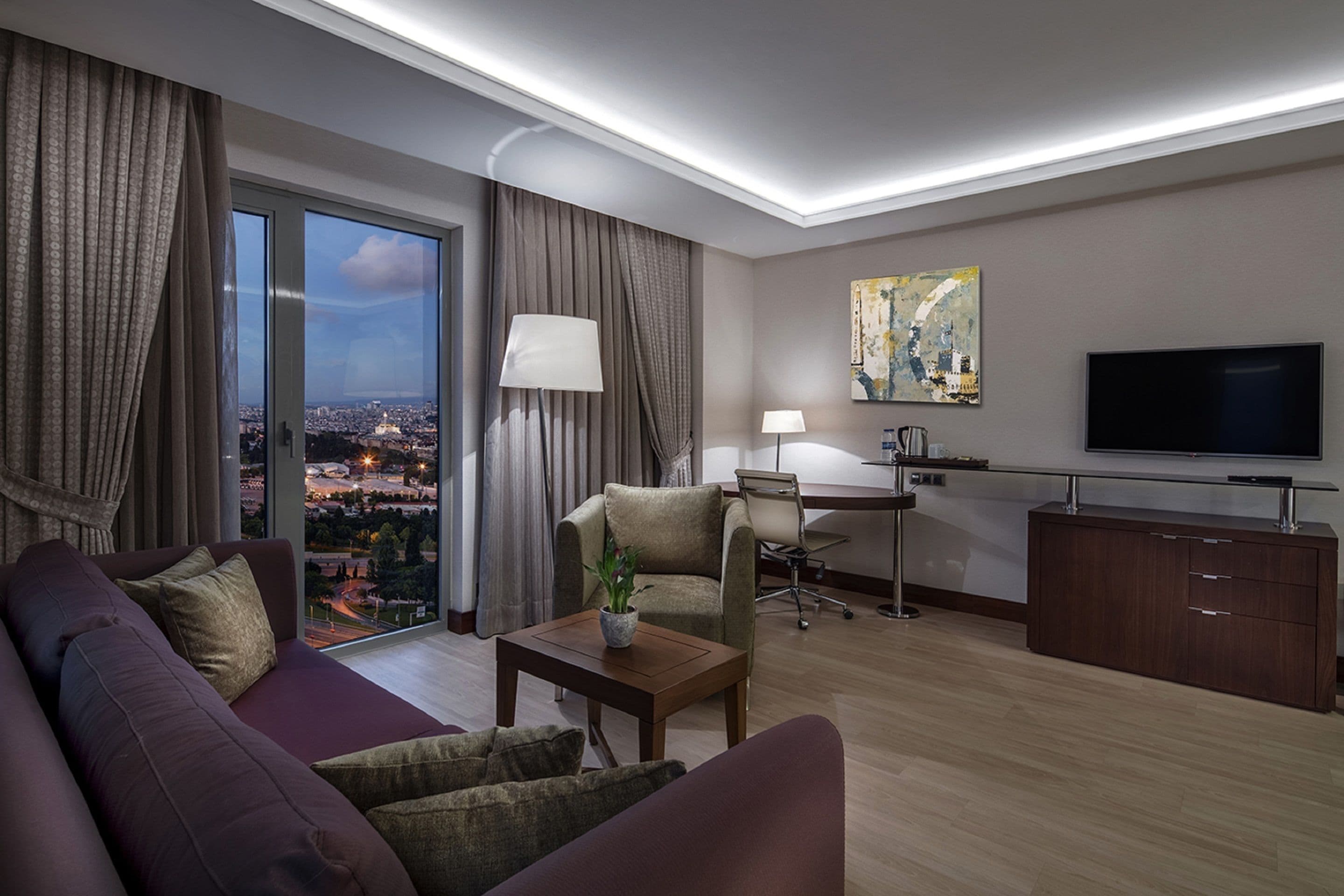 Ein modernes Hotelzimmer mit einer großen Couch, einem kleinen Tisch mit Pflanze, einem Schreibtisch mit Stuhl, einem Fernseher und Blick auf die Stadt durch das Fenster.