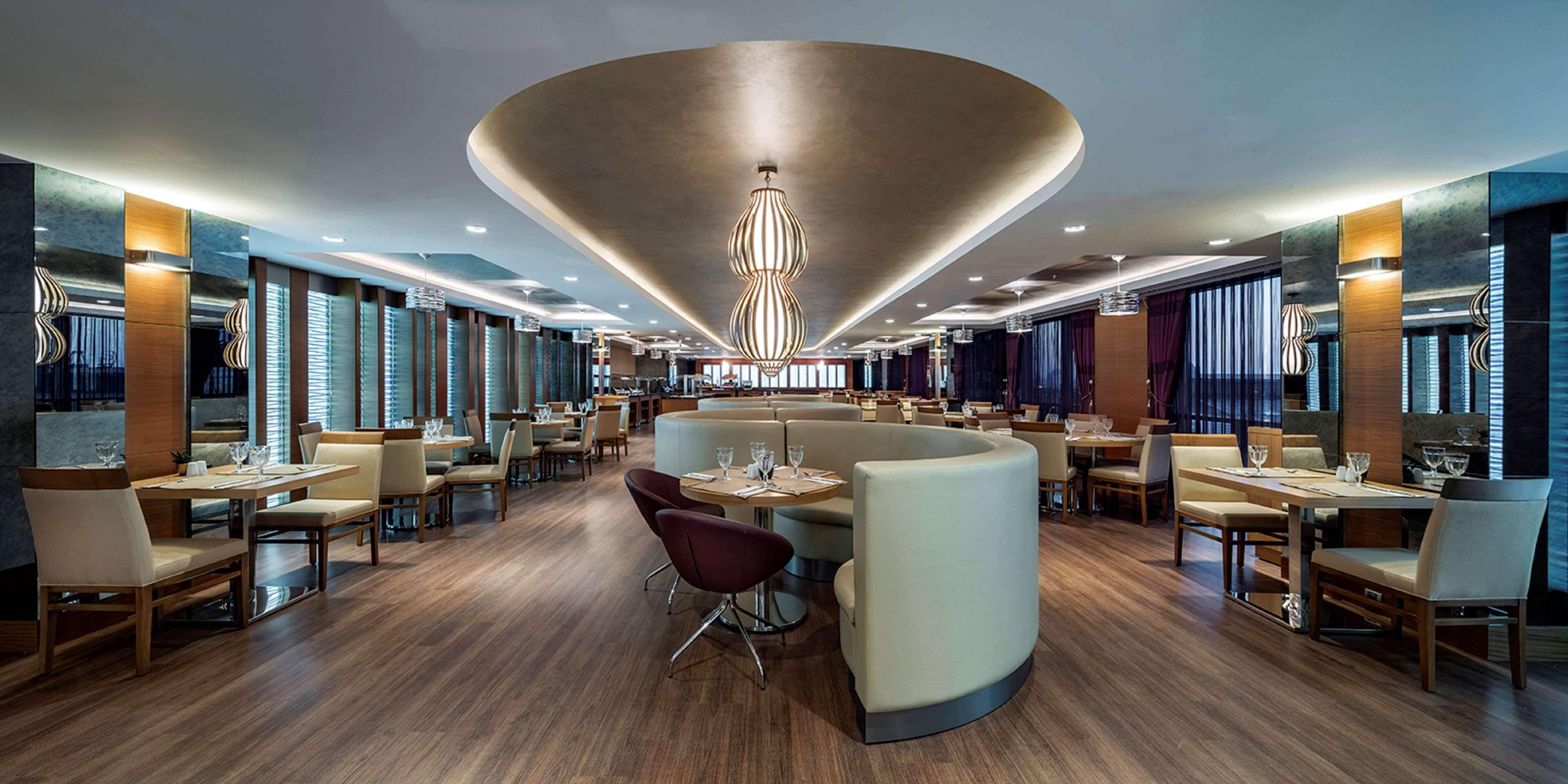 A modern restaurant with a central seating area, multiple dining tables, and a distinctive ceiling design.