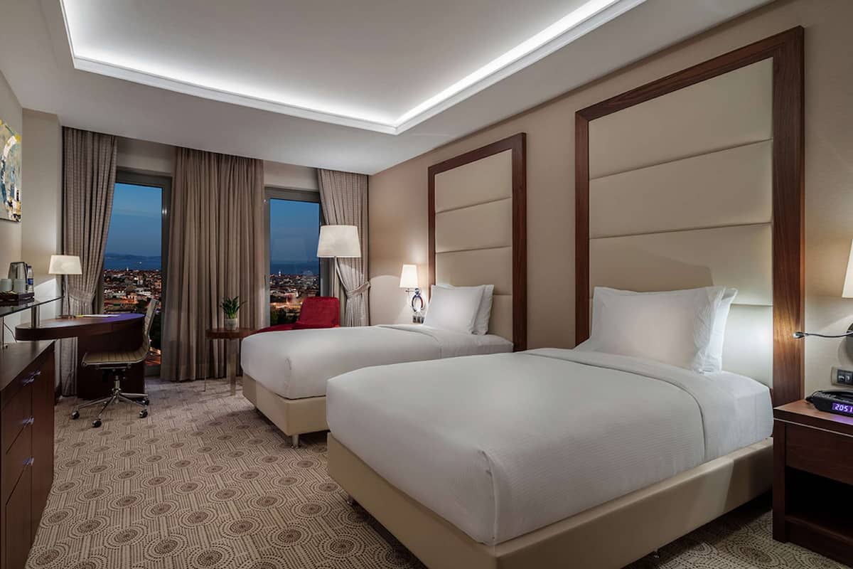 Quarto de hotel com duas camas, uma escrivaninha, televisão e vista para a cidade.