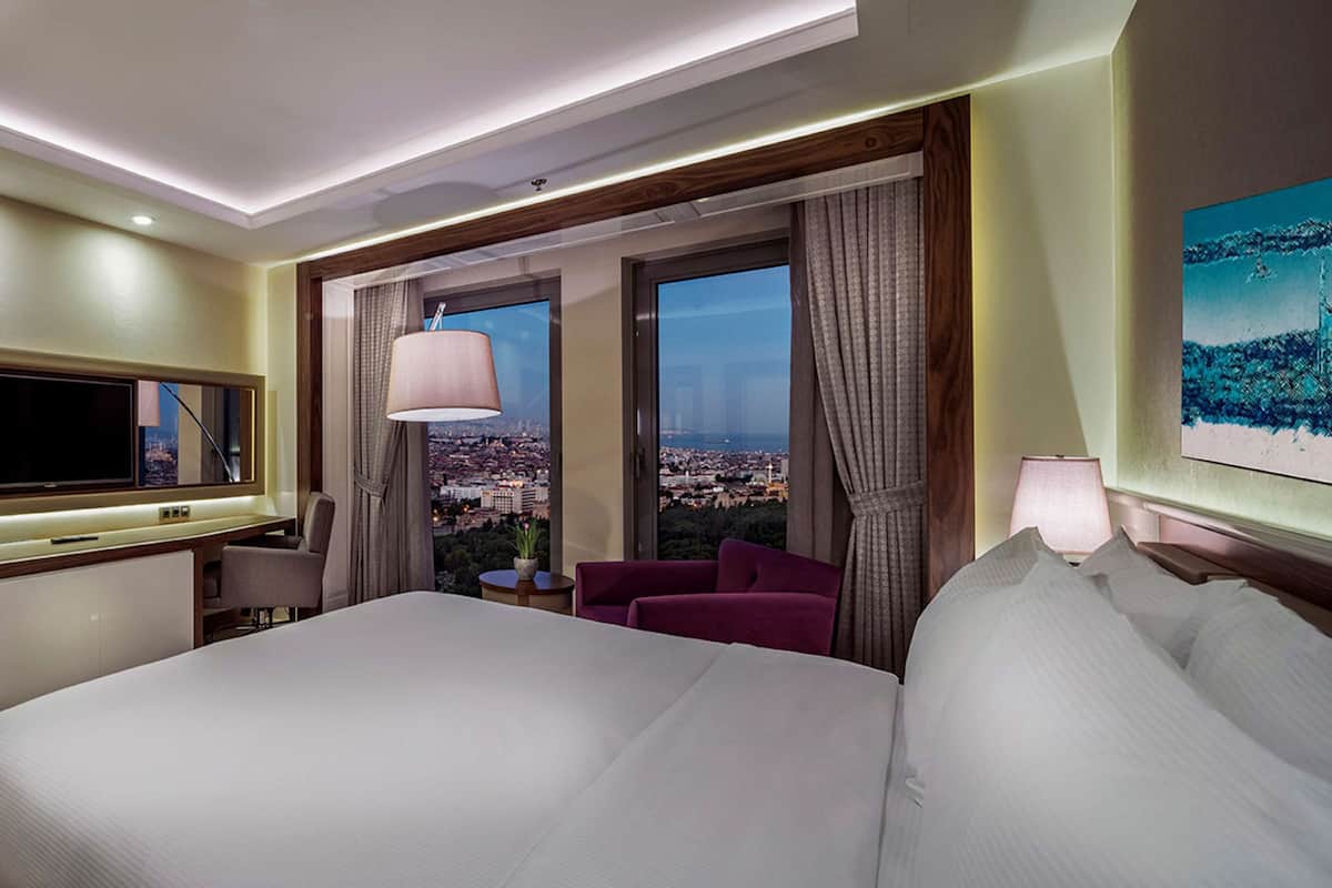 Quarto de hotel com uma cama grande, uma escrivaninha com TV de tela plana, área de estar com cadeiras e vista para o horizonte urbano através de amplas janelas.