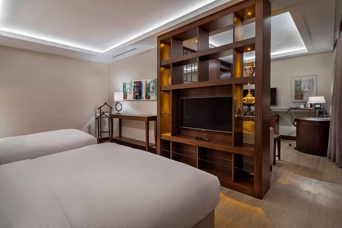 Quarto de hotel com duas camas, televisão, escrivaninha e um pequeno espaço para sentar.