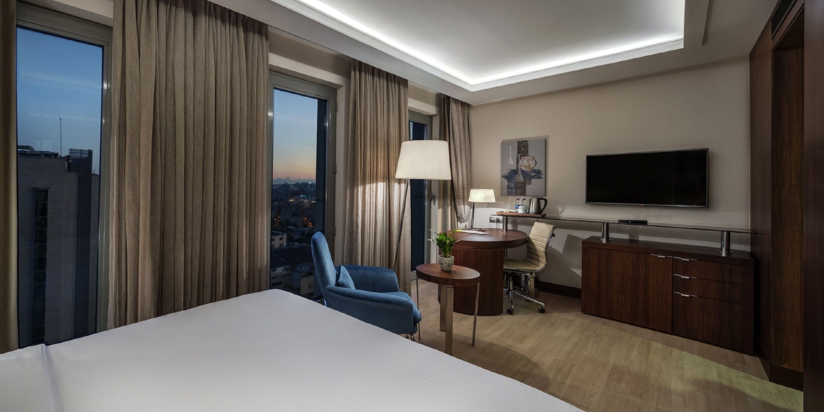 A hotel room with a large bed, a desk with a lamp, a chair, a TV, and a view of the city.