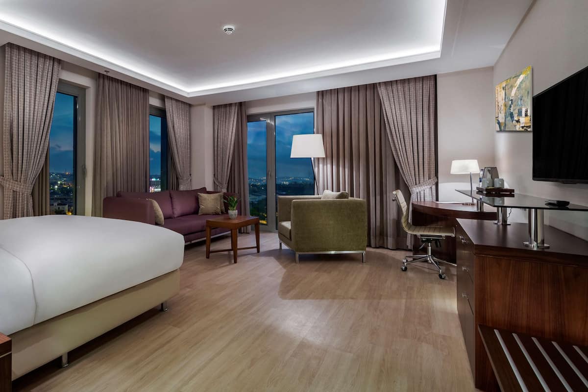 Quarto de hotel com uma cama grande, uma escrivaninha, uma cadeira, uma TV e vista para a cidade à noite.