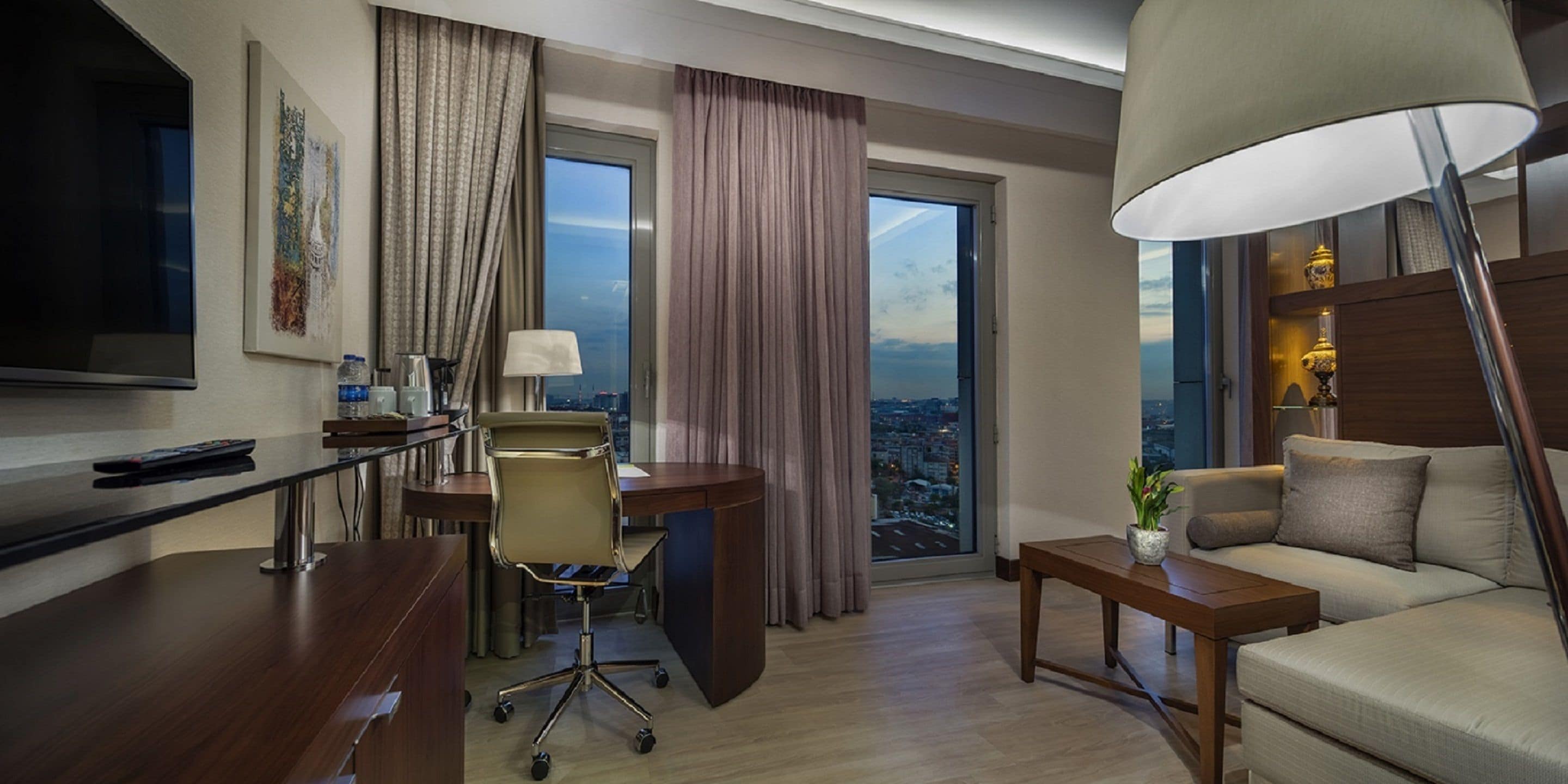 A modern hotel room with a desk, chair, sofa, and a large window offering a city view.