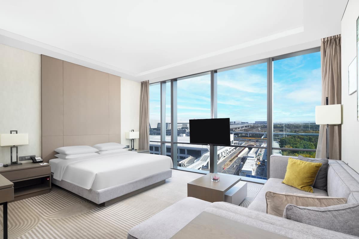 A modern hotel room with a large bed, a sofa, a TV, and a city view.