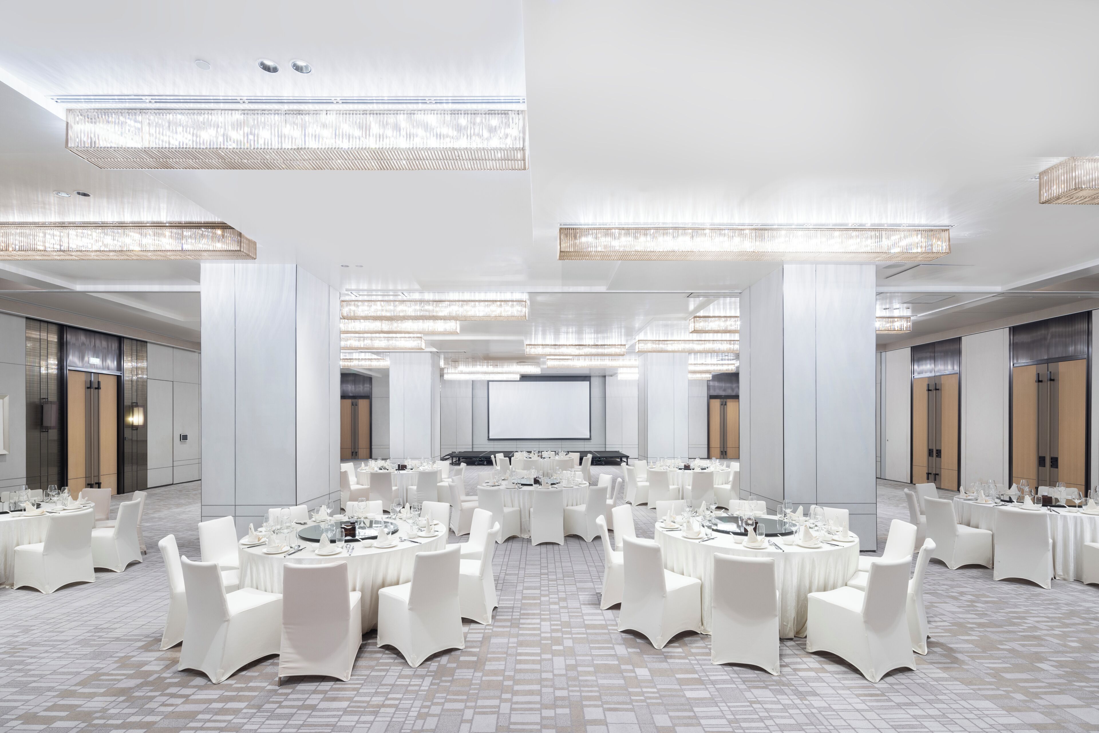 Une grande salle de conférence bien éclairée, avec des tables rondes, des chaises blanches et un écran de projection à l’avant.