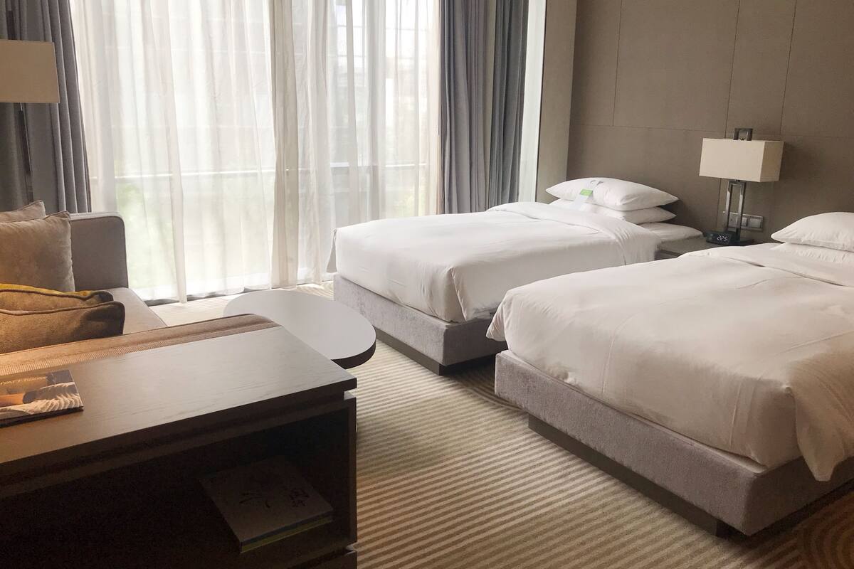 A hotel room with two beds, a desk, a chair, a lamp, and a window with curtains.
