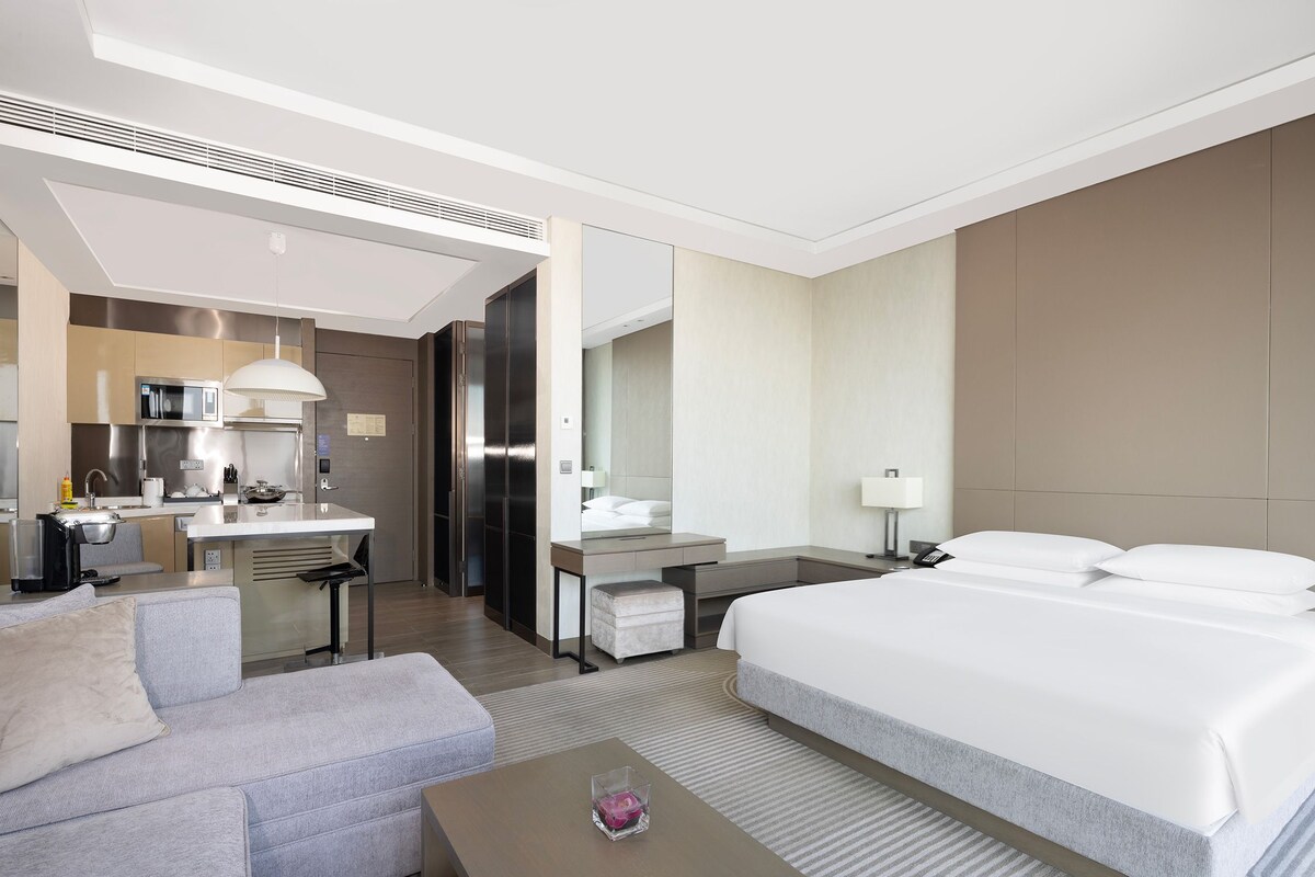 A modern hotel room with a bed, a sofa, a desk, and a kitchenette.