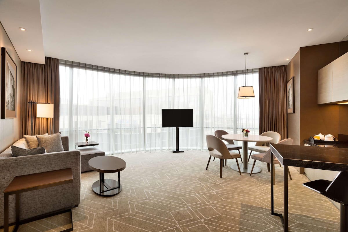 A modern hotel room with a sofa, a dining table, chairs, a TV, and a large window with curtains.
