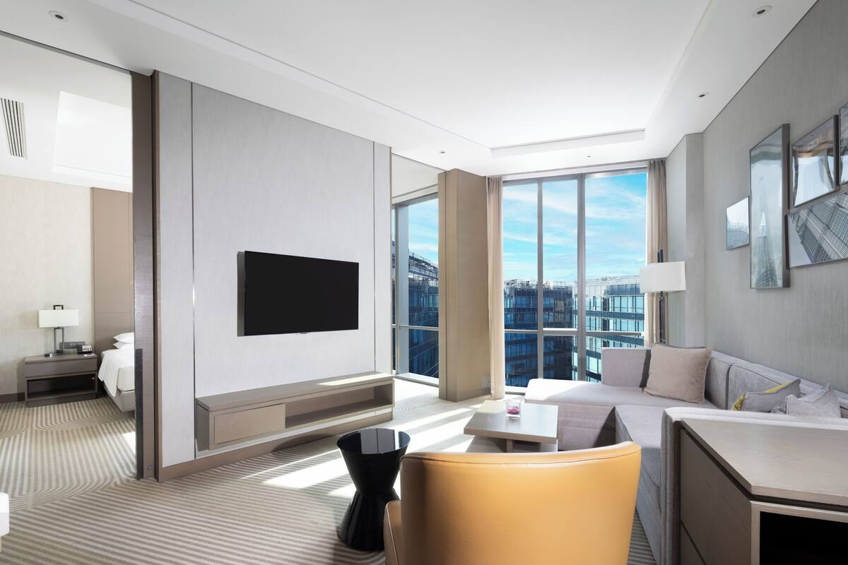 A modern hotel room with a large window, a flat-screen TV, a sofa, a dining table, and a chair.