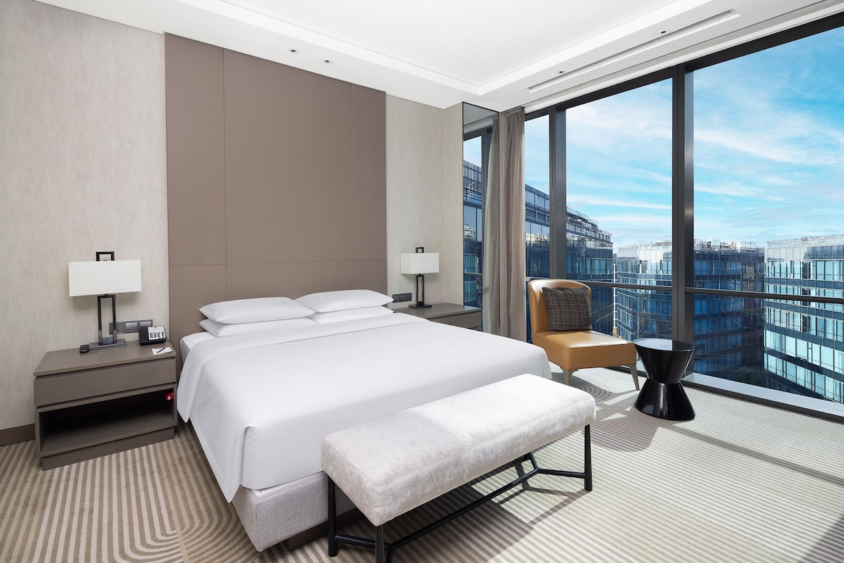 A modern hotel room with a large bed, bedside tables, a chair, and a view of the cityscape through large windows.
