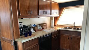 A compact kitchen with wooden cabinets, a dishwasher, a coffee maker, and a window with blinds. - Southern Comfort Yacht (Charleston)