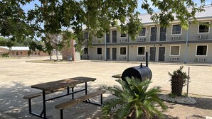 A picnic table with benches, a barbecue grill, and a palm tree in a courtyard with a building in the background.