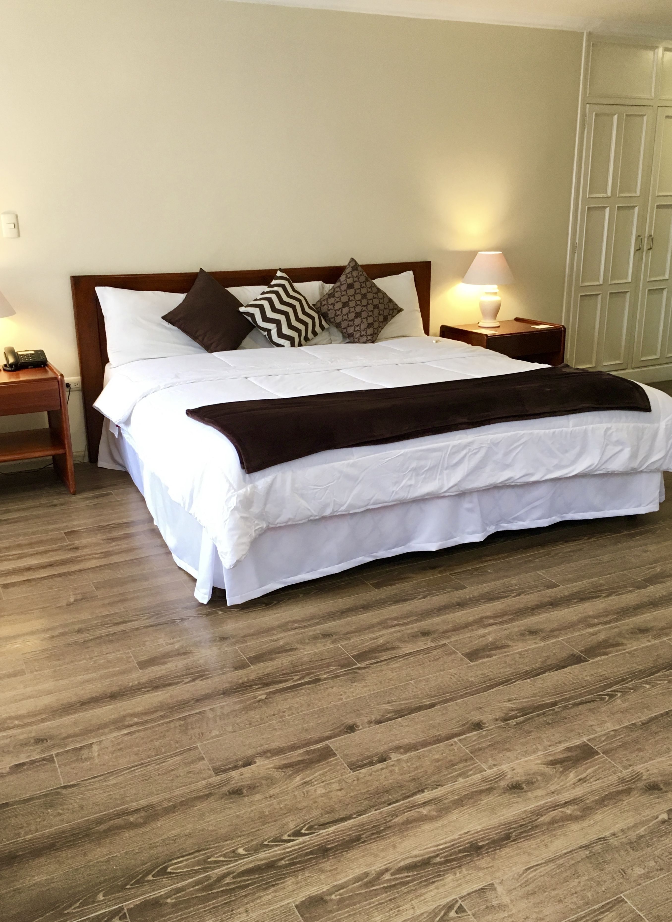 A neatly made bed with white linens and a brown throw blanket, flanked by two bedside tables with lamps, in a room with wooden flooring.