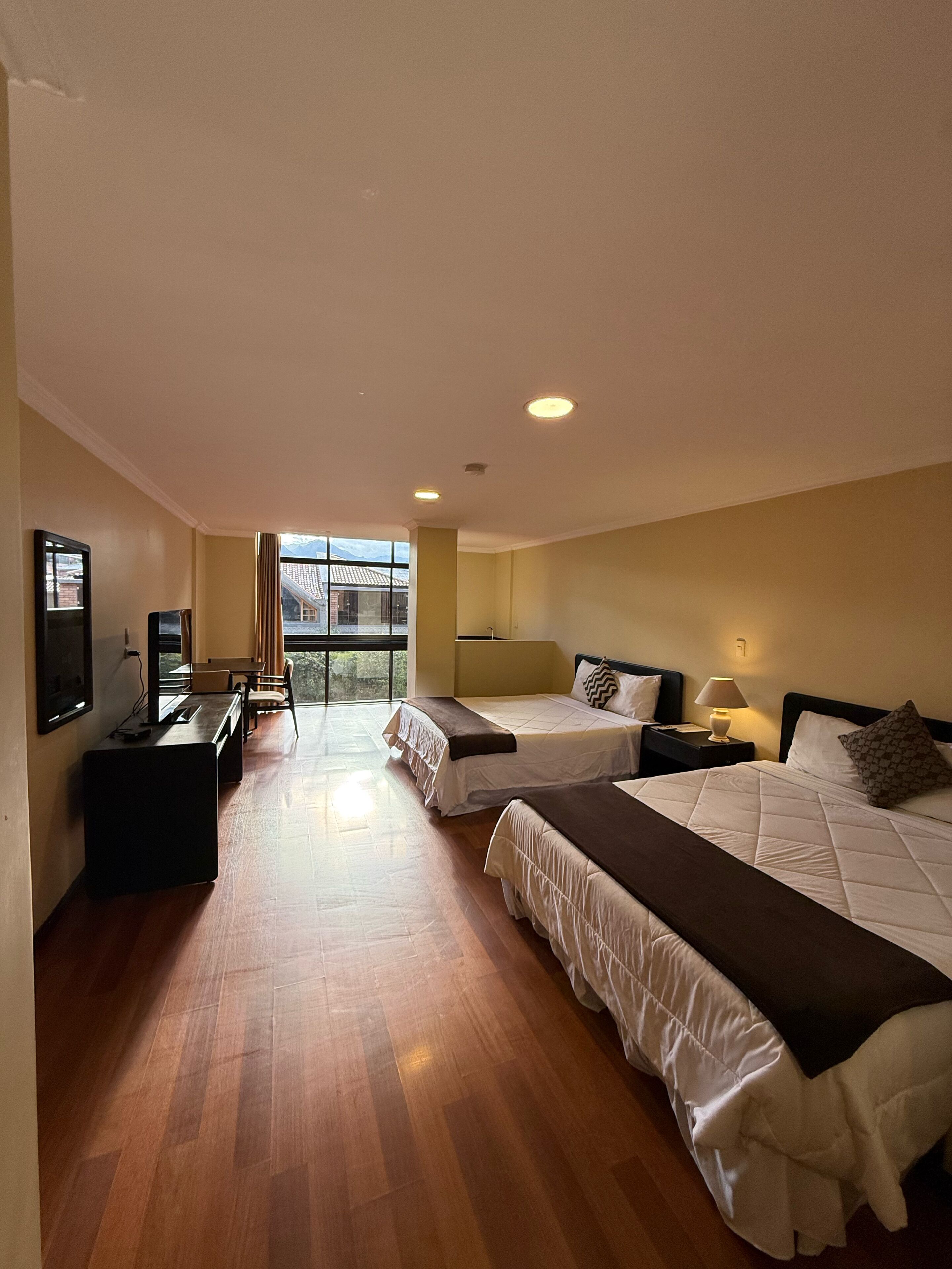 A hotel room with two beds, a desk, a chair, a TV, and a large window.