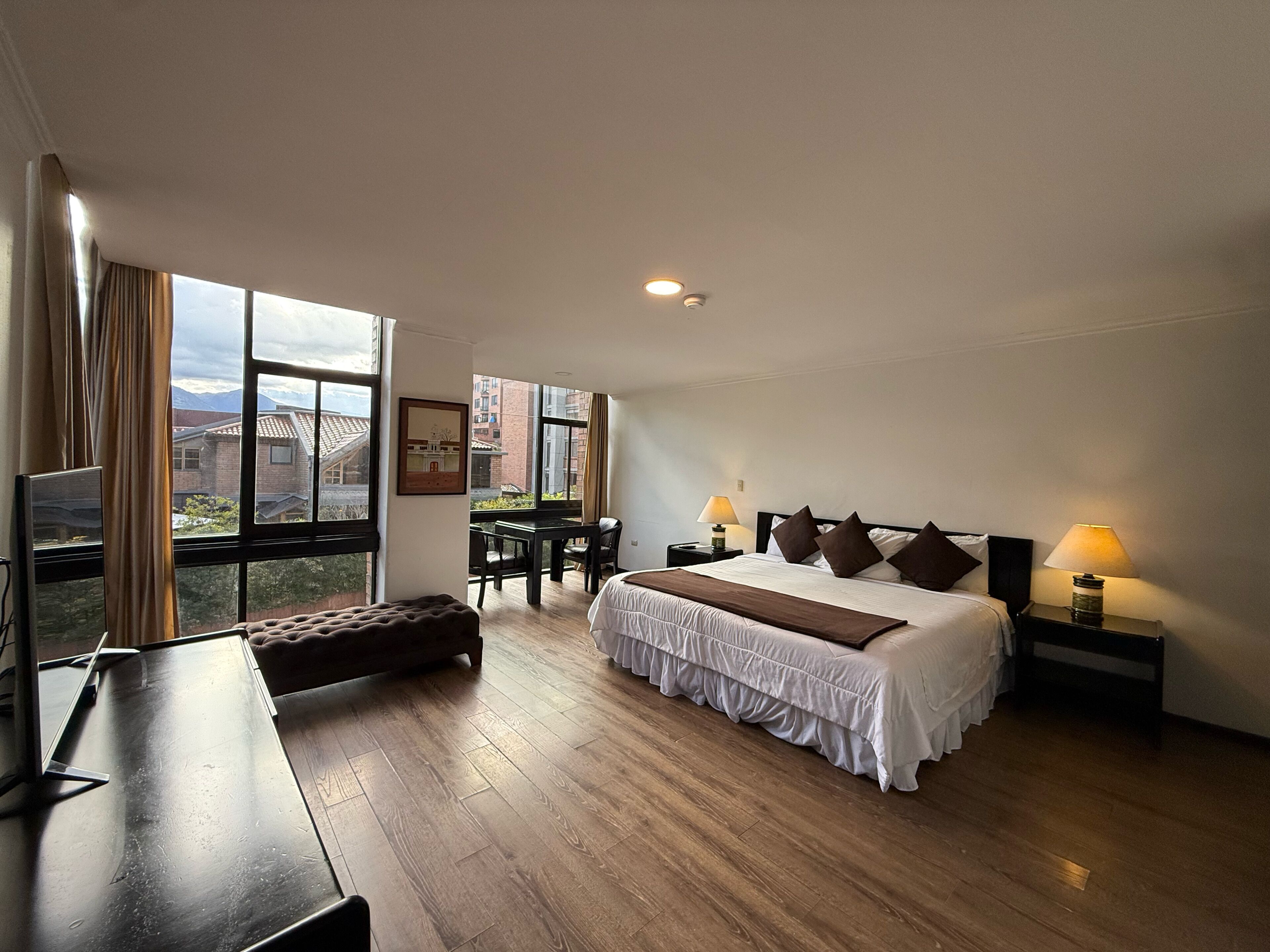 Een hotelkamer met een groot bed, nachtlampjes, een bureau en een raam met uitzicht op gebouwen en bergen.