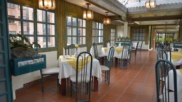Un restaurante con varias mesas y sillas, ventanas amplias y lámparas colgantes.