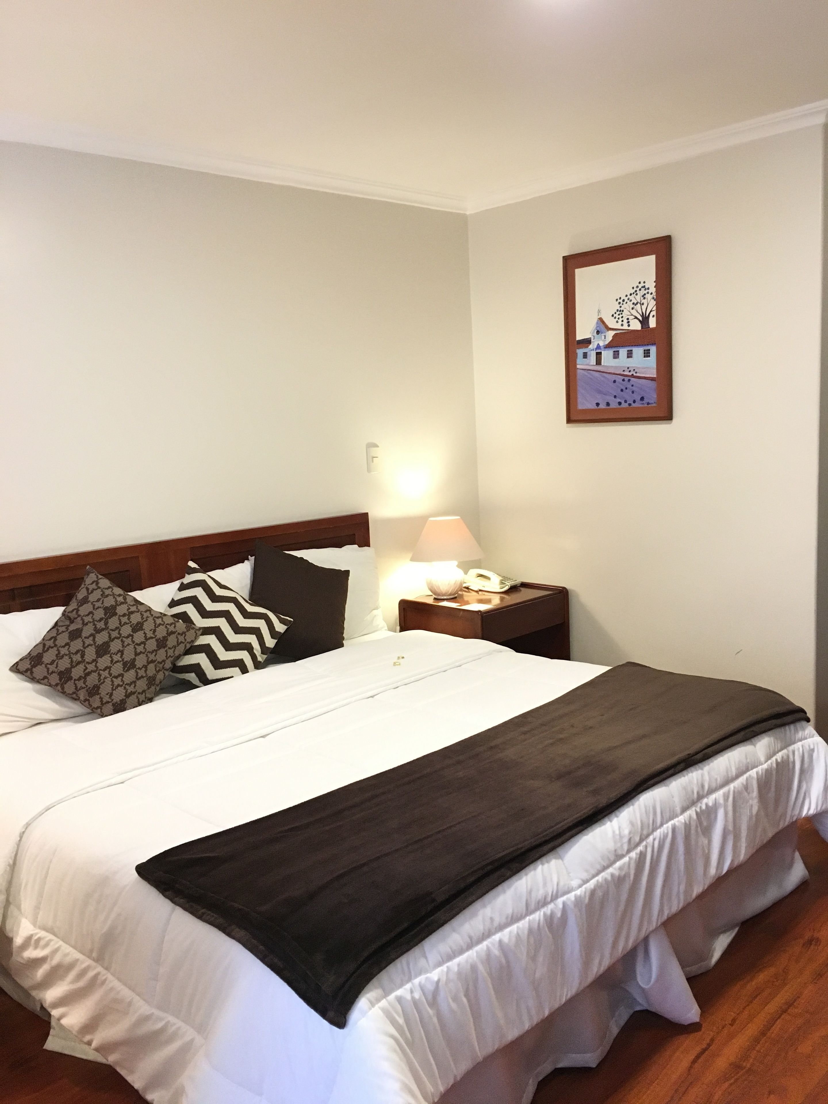 Una cama bien hecha con un edredón oscuro, dos almohadas estampadas, una lámpara de noche y un cuadro en la pared.