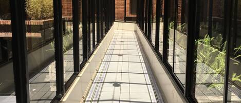 A covered walkway with glass walls and a tiled floor, leading to a brick building.