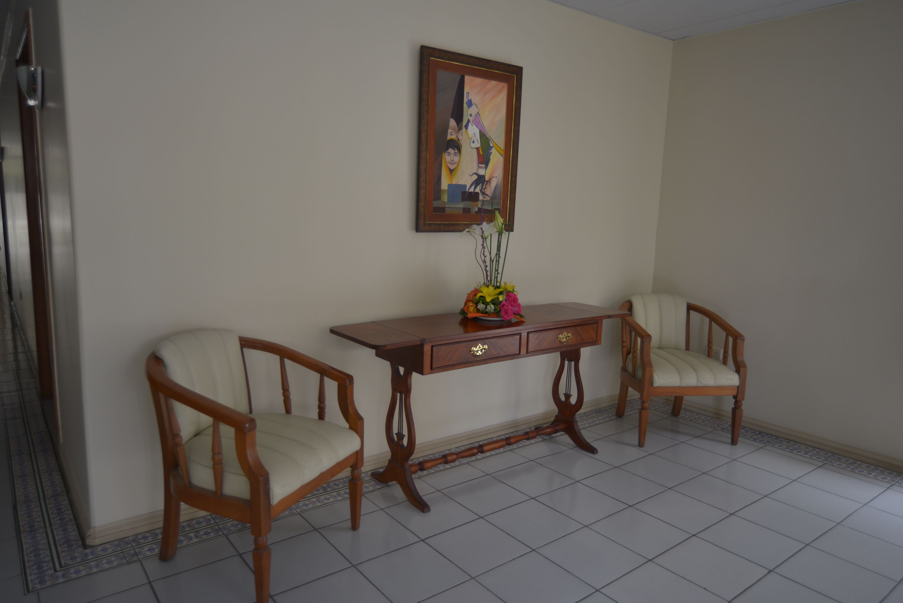 A room with two wooden chairs, a wooden table with drawers, a vase with flowers, and a framed picture on the wall.