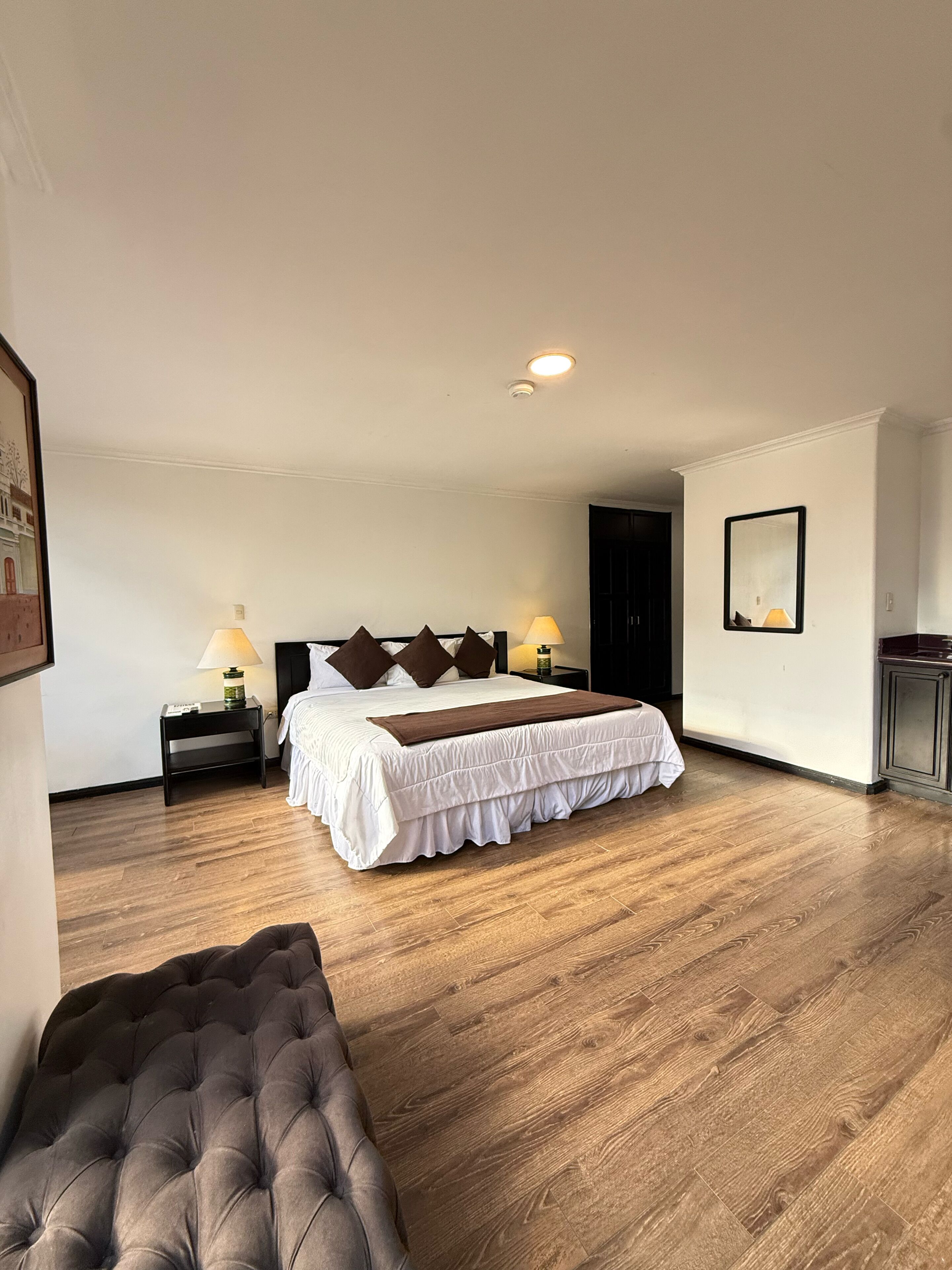 A bedroom with a large bed, two bedside tables with lamps, a dark-colored tufted chair, and a framed picture on the wall.