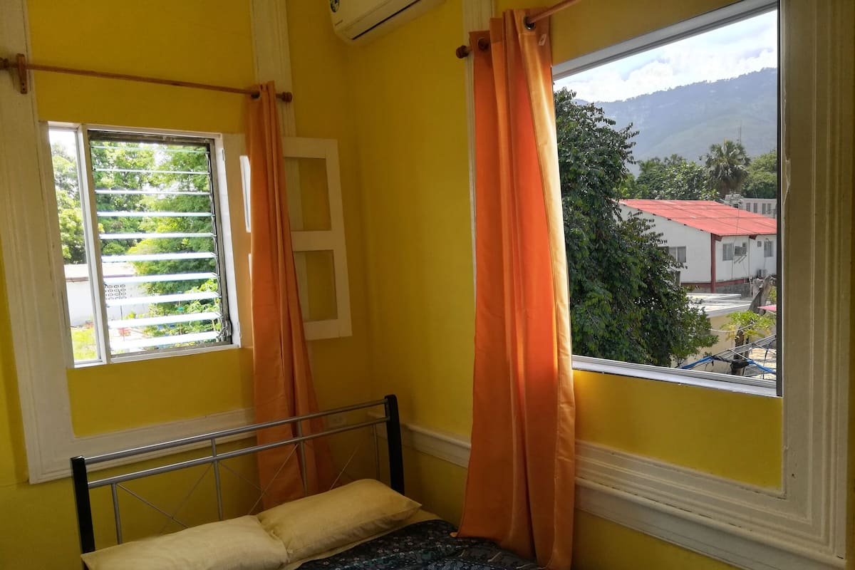 Une pièce avec un lit, une fenêtre avec des rideaux et une vue sur les bâtiments et les arbres à l’extérieur.