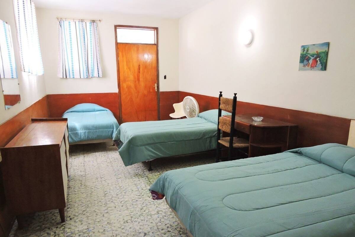 Une chambre avec quatre lits simples, un bureau en bois, une chaise, une petite table et un ventilateur.