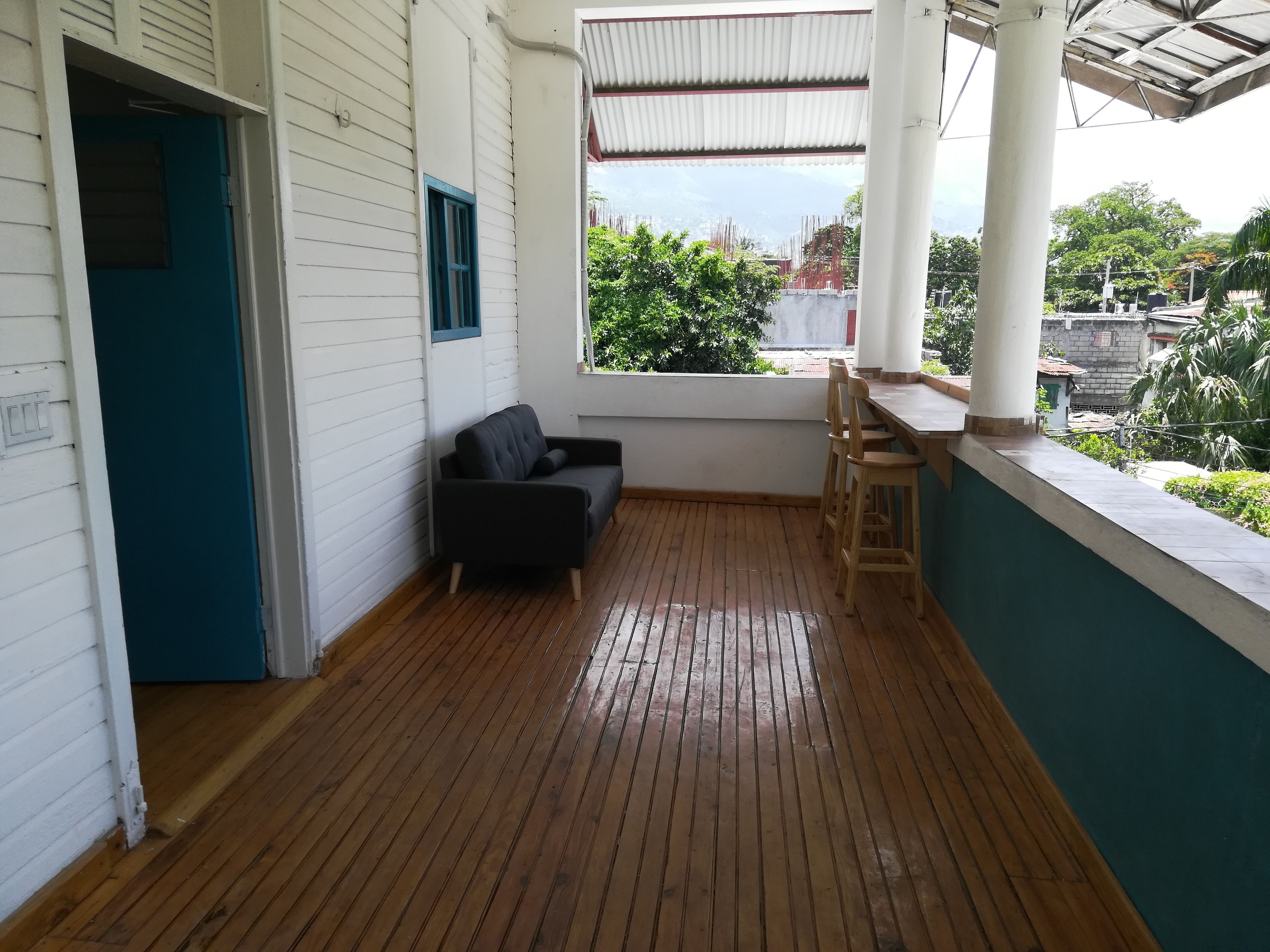 Una habitación con piso de madera, sofá negro, ventana con vista a zonas verdes y pared blanca con puerta azul.