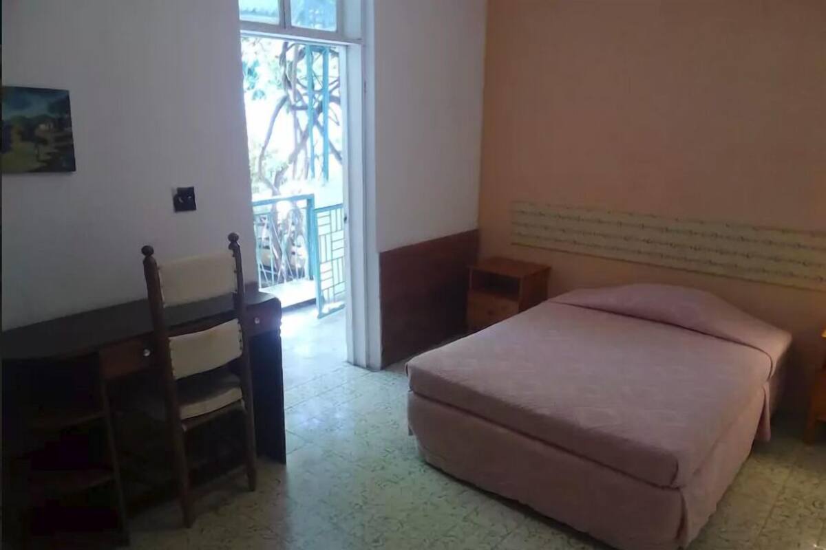 Une chambre à coucher avec un lit, un bureau et une chaise. Il y a une fenêtre donnant sur des arbres et un balcon.