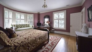 A bedroom with a large bed, a desk, and a chair. There are two windows with shutters and a chandelier. - The Peter Herdic Inn (Williamsport)