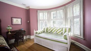 A room with a curved bay window, a green-striped sofa bed, a wooden desk with a lamp, and a framed picture on the wall.