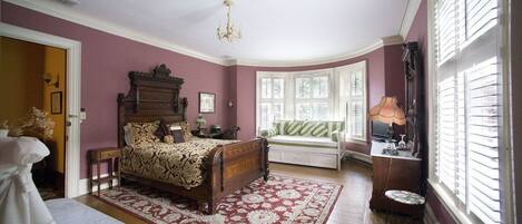 A bedroom with a wooden bed, a bench, a table, and a window with blinds.
