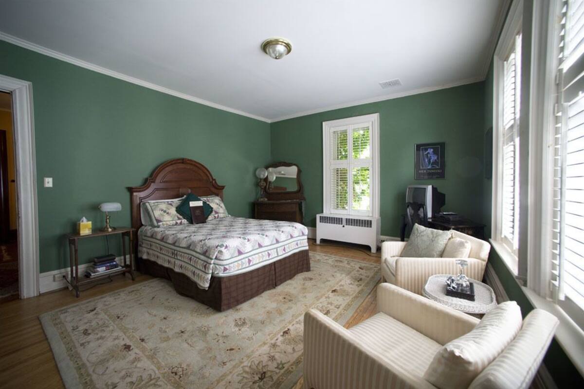 A bedroom with a large bed, a chair, a nightstand, a window with blinds, and a radiator.