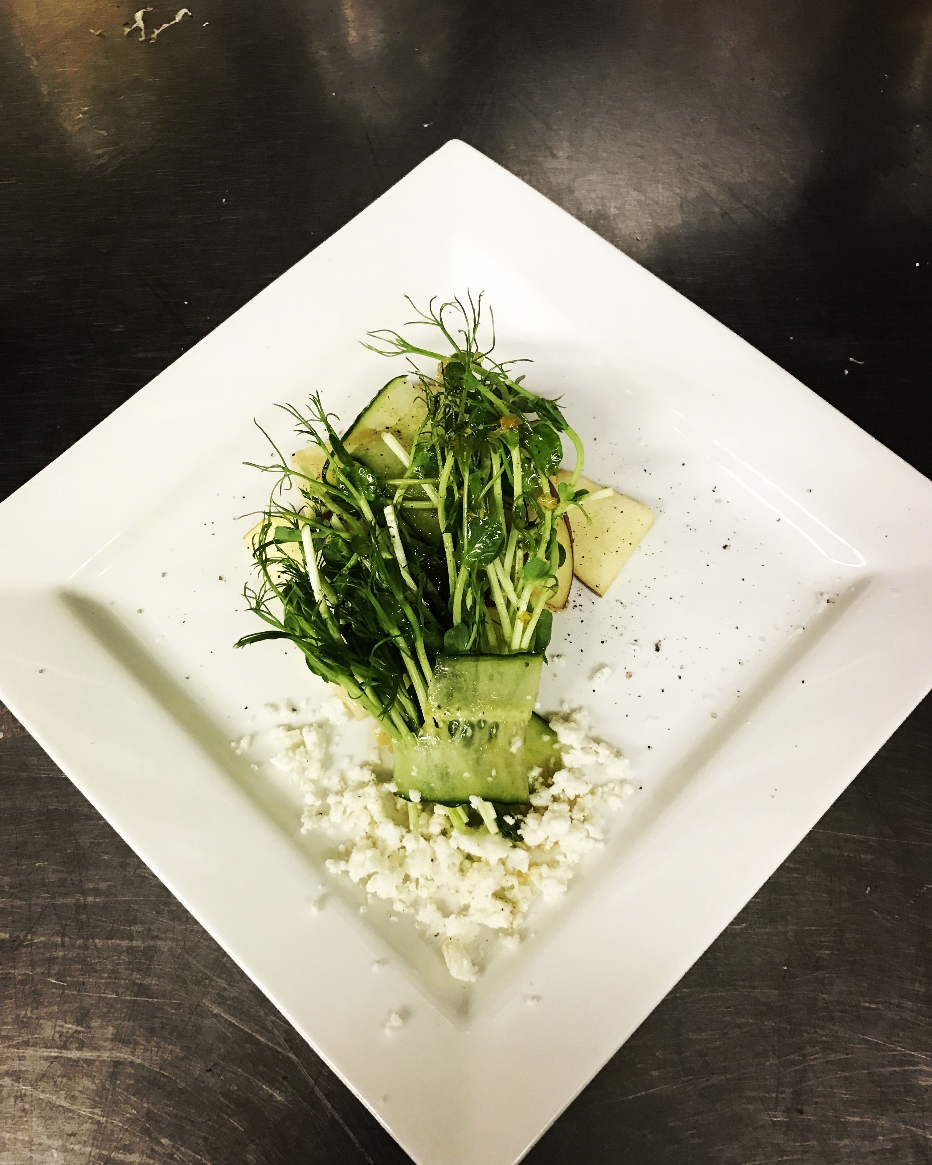 A white square plate with a salad consisting of cucumber slices, greens, and crumbled cheese on a dark surface.