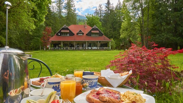A breakfast table with various foods and drinks, a kettle, and a view of a large house surrounded by trees. - Wedgwood Manor and Glamping Retreat (Crawford Bay)