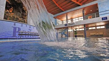 Una piscina con caída de agua, un cartel que dice "ESTAC" y un edificio de varios niveles con balcones.
