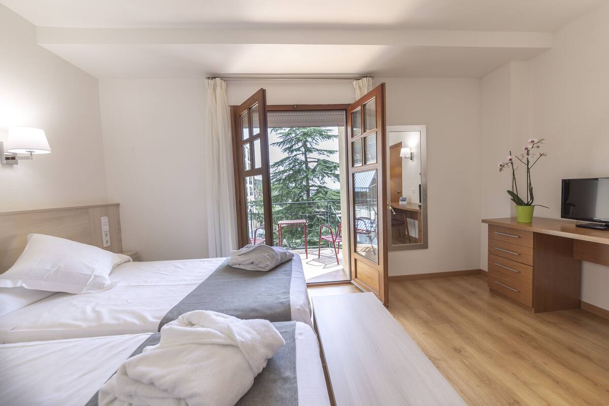 A hotel room with two beds, a desk, a TV, and a balcony with a view of trees.