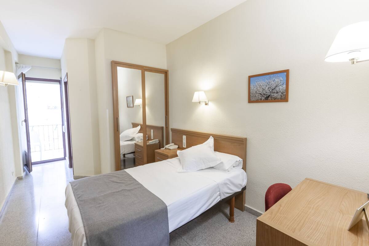 A hotel room with a bed, bedside table, chair, desk, and a mirror.