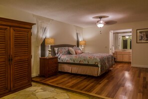 Quarto com piso de madeira, cama com roupa de cama florida, duas luminárias de cabeceira, guarda-roupa de madeira e ventilador de teto.