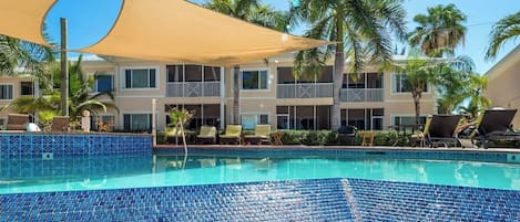 Ein Hotelpoolbereich mit einem blau gefliesten Pool, Liegestühlen, Palmen und einem Gebäude mit Balkonen.
