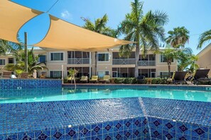 Ein Hotelpoolbereich mit einem blau gefliesten Pool, Liegestühlen, Palmen und einem Gebäude mit Balkonen.