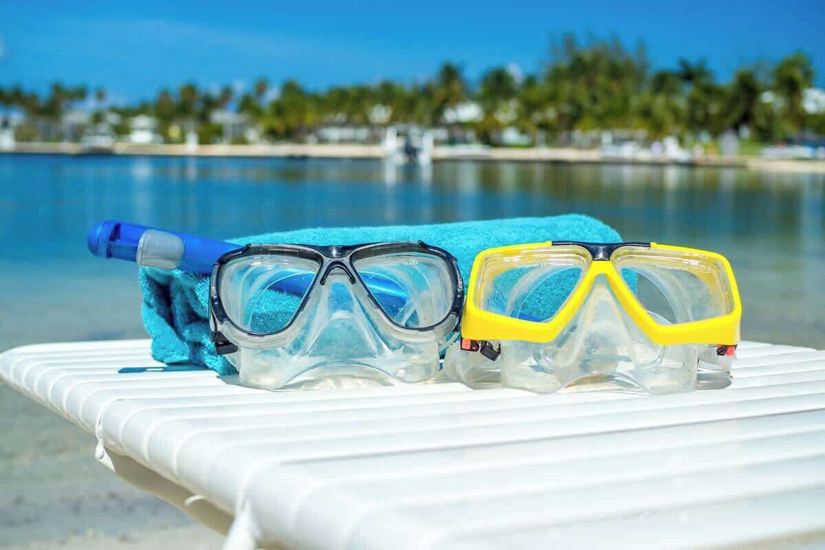 Snorkeling gear and a blue towel on a white lounge chair by a pool with a clear view of palm trees and a calm sea.
